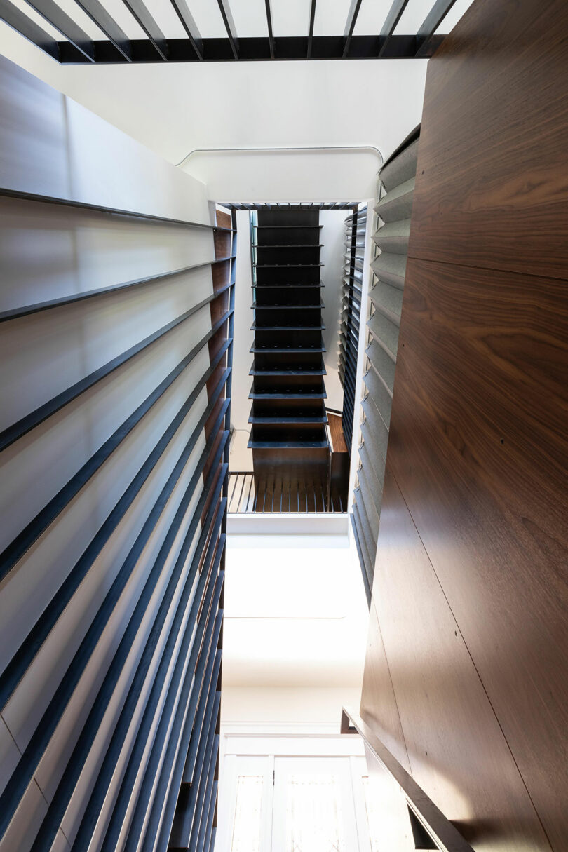 View from the bottom of a modern staircase with dark, measured steps, wooden paneling, and metal railings, looking upwards to a bright skylight.