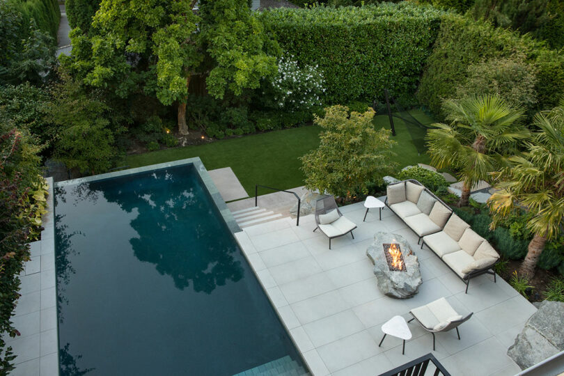 Aerial view of a modern backyard featuring a rectangular swimming pool, patio with a fire pit, outdoor furniture, and a lush garden bordered by tall hedges.