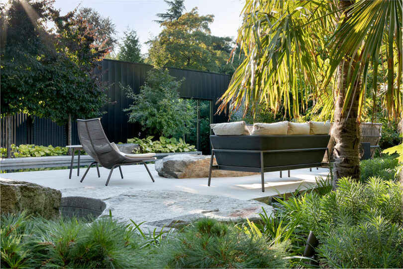 Modern garden featuring lush greenery, a black sofa with cushions, a lounge chair, and large rocks, surrounded by a black fence.