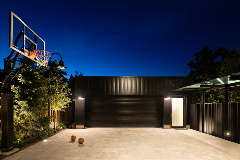 A modern outdoor home basketball court at night, featuring a hoop, paved area, and illuminated garage and trees.