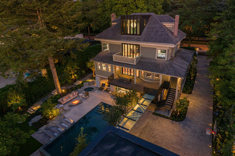 Aerial view of a large three-story house at dusk, featuring illuminated windows, and a swimming pool.