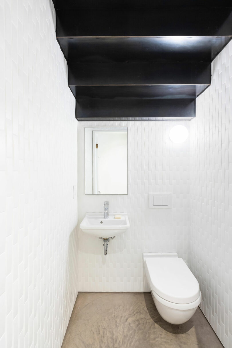 A small, modern bathroom designed with white textured walls, a wall-mounted sink, toilet, and a black ceiling. A single mirror and overhead light fixture are visible.