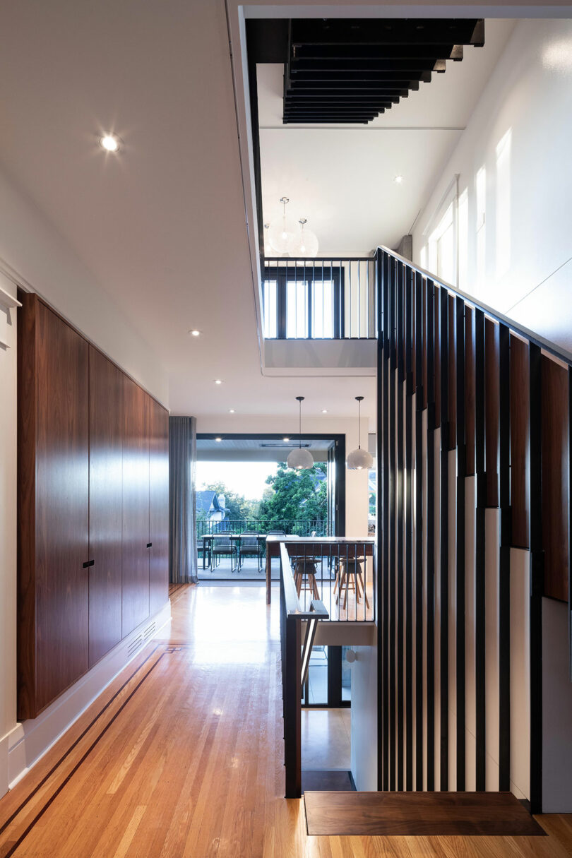 Modern home interior featuring a hallway with wooden floors and walls, a staircase with vertical black bars, and a view into a well-lit, open room with large windows.
