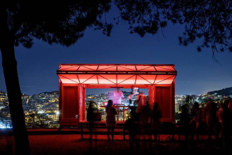 People gathered around a lit-up pavilion at night with a cityscape backdrop.