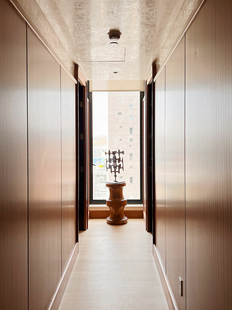 Narrow hallway at with wooden doors and floors, leading to a window displaying a vase on a pedestal; sunlight illuminates the space.