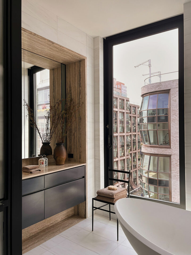 Modern bathroom with a large mirror, wooden vanity, freestanding tub, and open window showing city view.