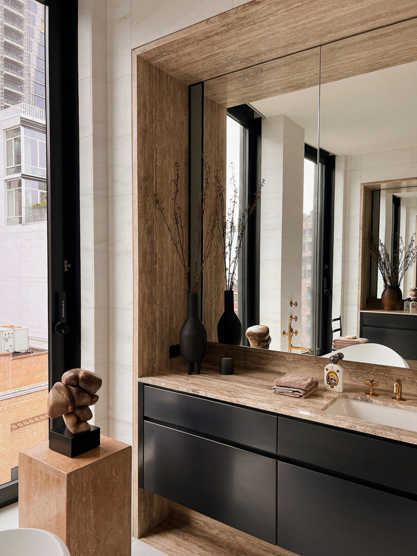 A modern bathroom vanity with black drawers, a large mirror, decorative vases, and a city view through windows.