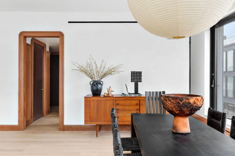 Modern dining room with a large black table, oversized paper pendant light, and wooden sideboard, featuring minimalist decor and large windows.