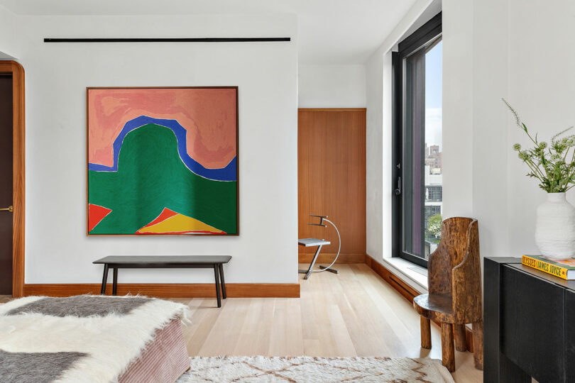 Modern living room with a large abstract painting, minimalist furniture, and a view of through floor-to-ceiling windows.