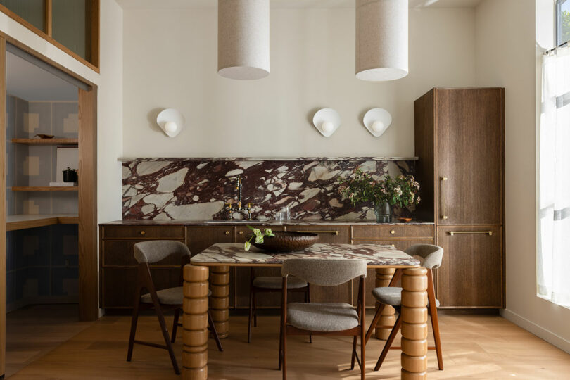 Modern dining room with a marble backsplash, round wooden table, and unique cylindrical chairs, complemented by pendant lighting and wooden cabinets.