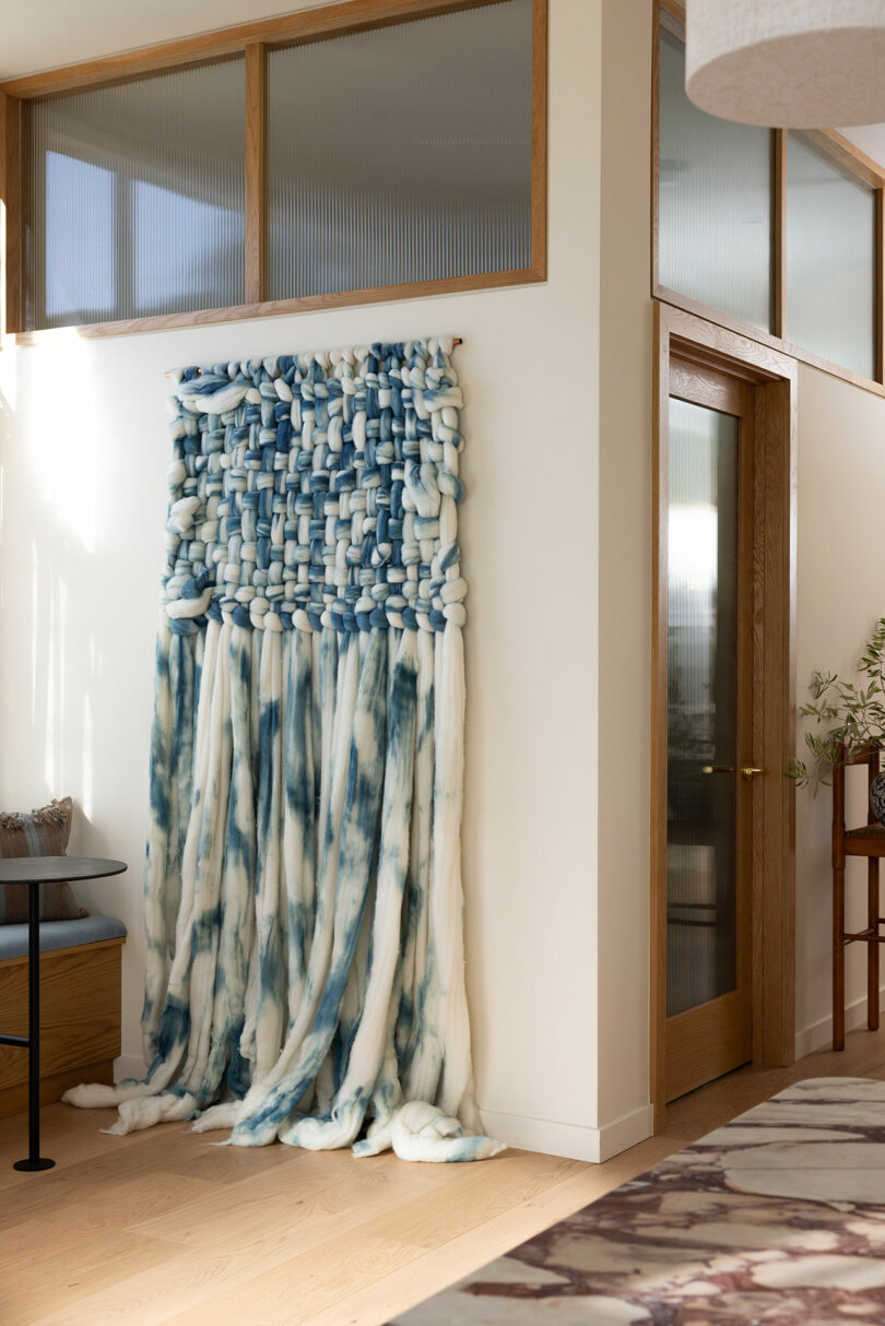 A large, textured wall hanging in shades of blue and white displayed in a modern room with wooden floors and glass doors.
