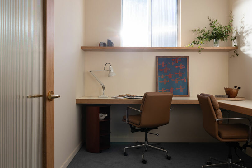 A small, well-lit office featuring two chairs, a desk with a lamp and documents, a shelving unit with a plant and framed art, and a frosted glass door.
