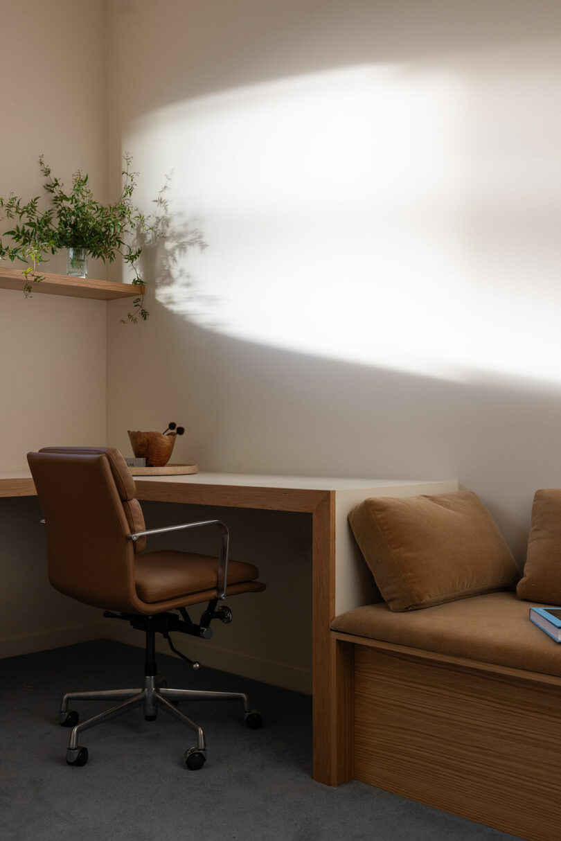 A cozy office corner featuring a wooden desk with a brown leather chair, a sofa with cushions, a houseplant on a shelf, and a striking light beam on the wall.