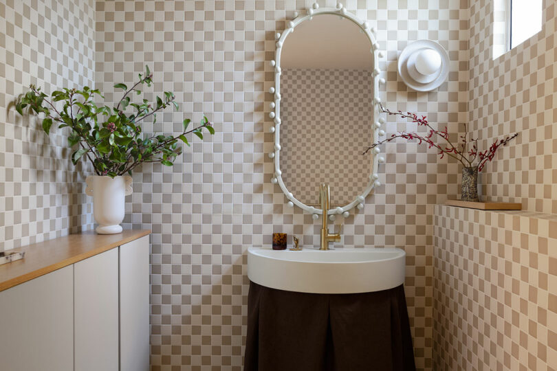 Modern bathroom with checkered tile walls, a round mirror with vanity lights, countertop sink, and decorative plants.