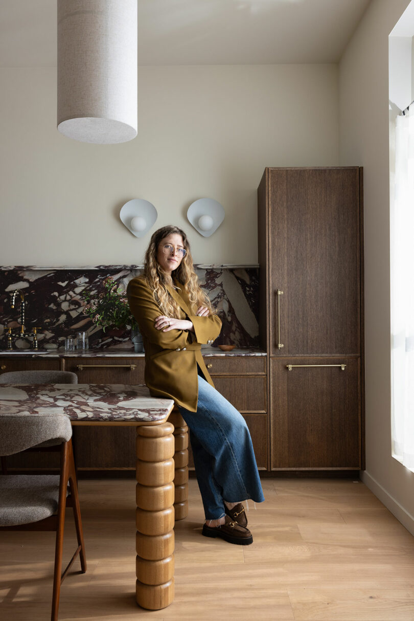 A woman in a gold blazer and jeans sits on a wooden stool in a stylish kitchen, leaning on a marble countertop.