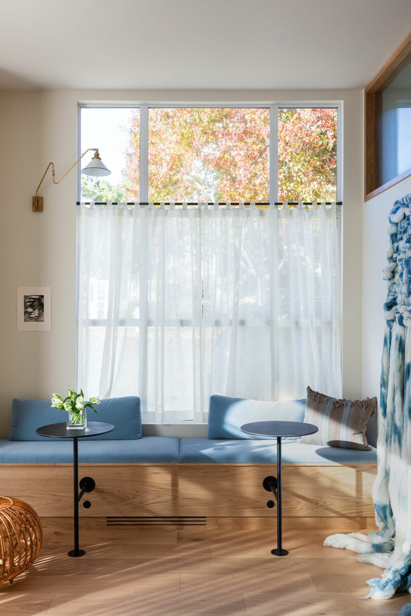 Bright living room with large window showing autumn trees, blue couch, two small tables, and sheer curtains.