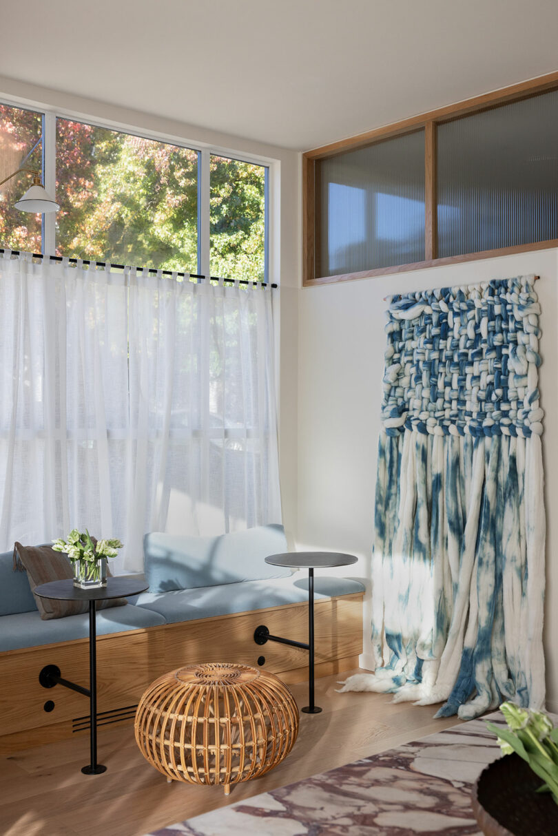 Modern living room with a blue sofa, woven circular coffee table, abstract fabric wall art, and large windows draped with sheer curtains.