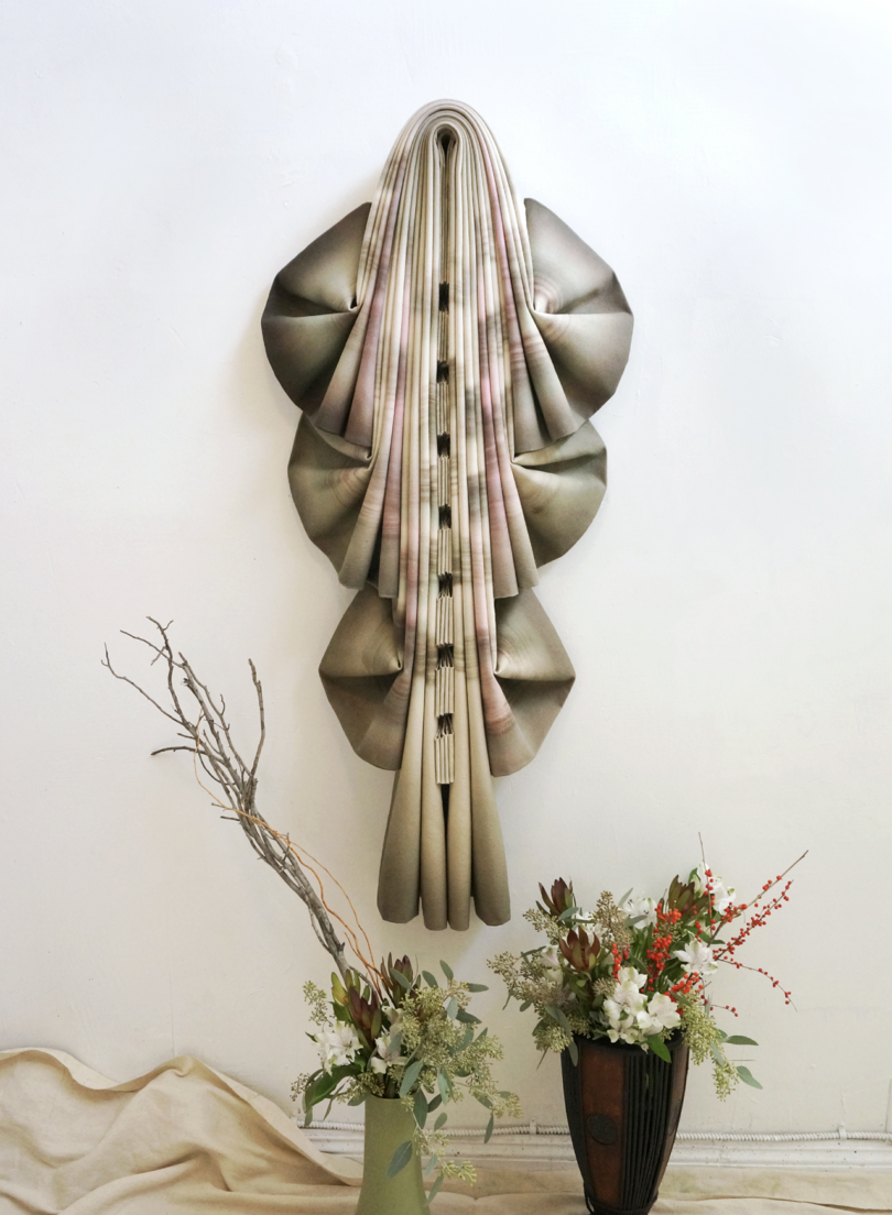 An artistic wall hanging resembling draped fabric accompanied by a branch and a floral arrangement in a vase.