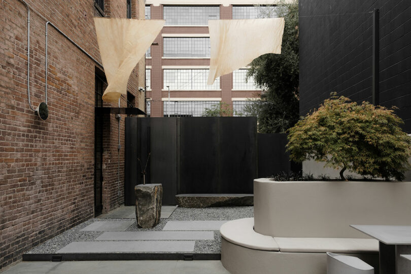 Modern urban courtyard with fabric shades, concrete seating, and a landscaped area against brick and dark panel walls.