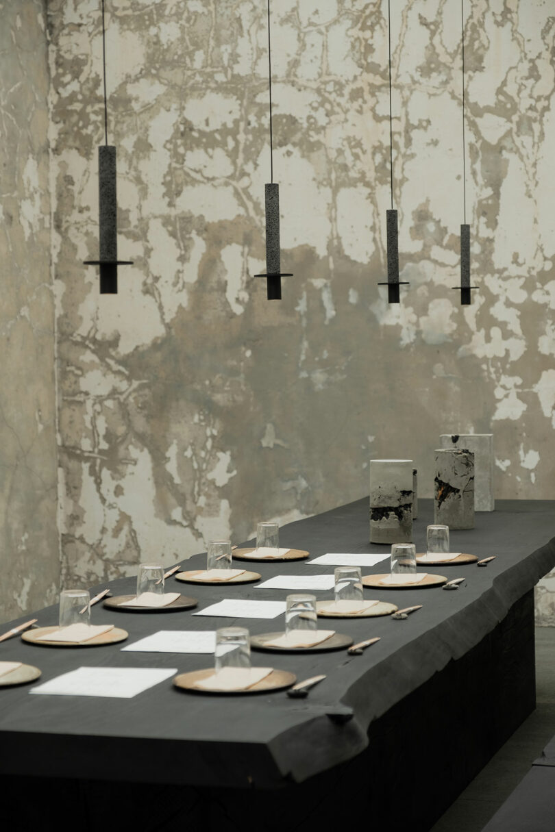 A minimalist dining setup with a long dark table, simple place settings, and cylindrical hanging lights, against a textured, neutral backdrop.
