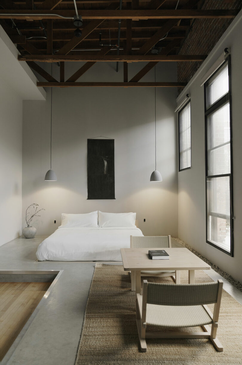 A minimalist bedroom featuring a white bedding against a light grey wall, with hanging pendant lights, exposed wooden beams, and a large window.