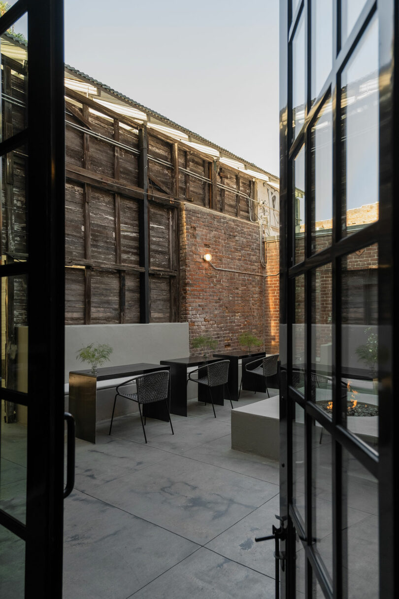 A modern patio with seating next to a historic brick building, viewed from indoors through glass doors.