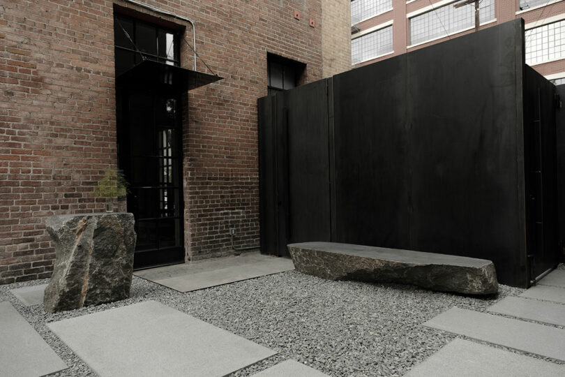 A minimalist urban courtyard featuring a large flat stone bench, gravel ground cover, and a dark, imposing privacy screen against a brick building.
