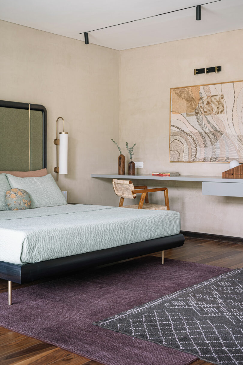 Modern bedroom with a minimalist design featuring a green and black bed, beige walls, abstract wall art, a wooden desk with a chair, and a purple rug.
