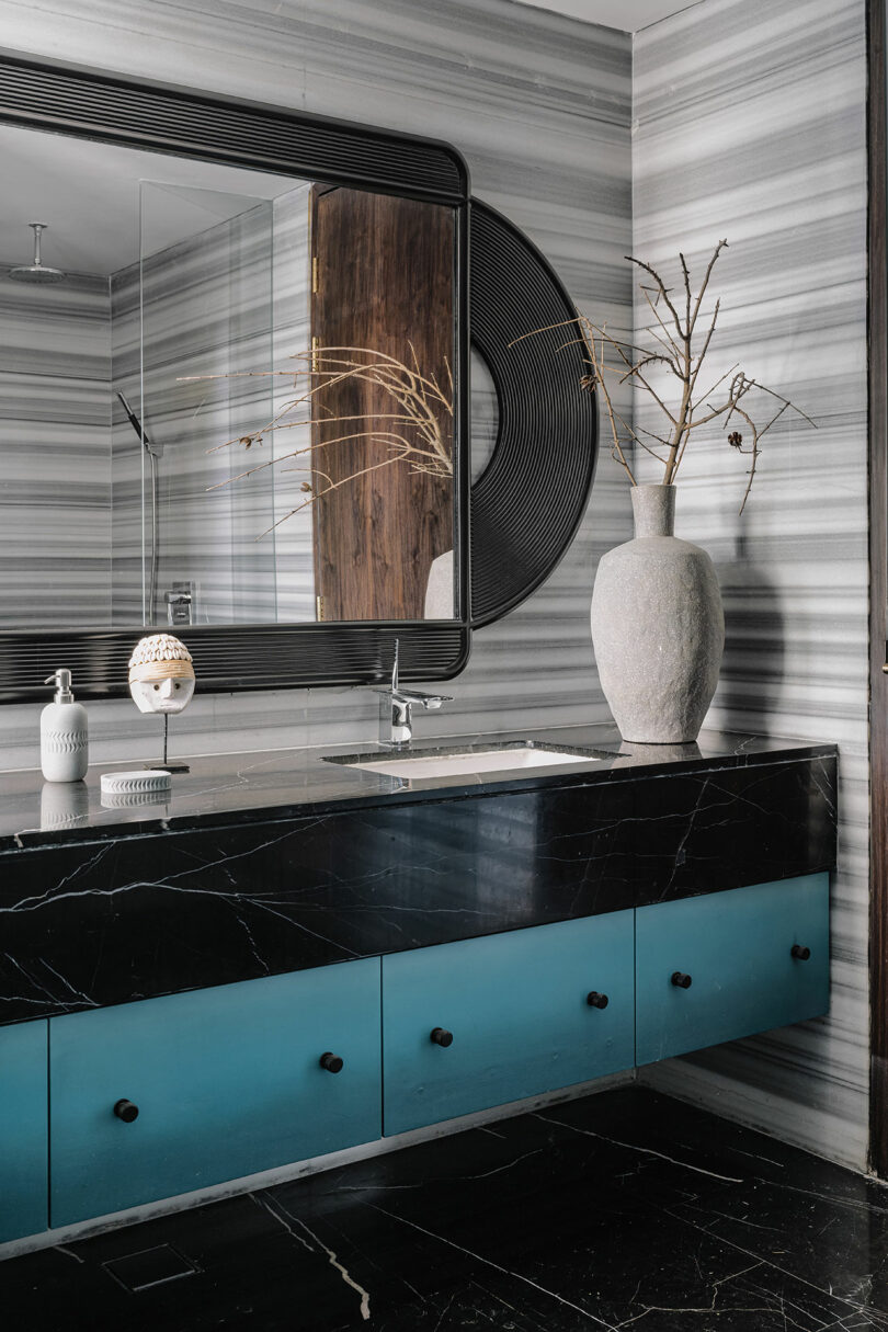 Modern bathroom with a teal vanity, black marble countertop, and a large mirror on a striped wall, decorated with a vase and branches.