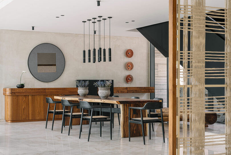 Modern dining room with wooden table and chairs, concrete walls, and artistic hanging lights. decor includes a circular mirror and wooden partition.