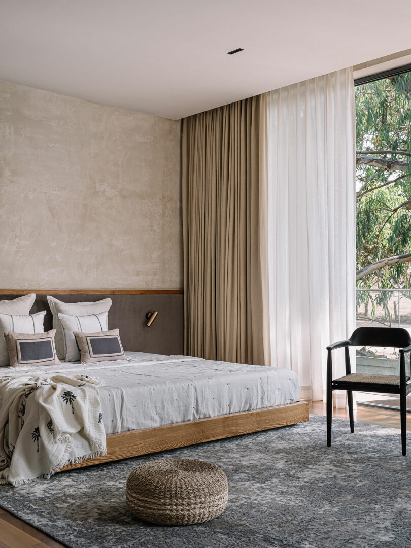 Modern minimalist bedroom with a low bed, beige walls, large window with sheer curtains, a dark chair, and a woven ottoman on a textured rug.