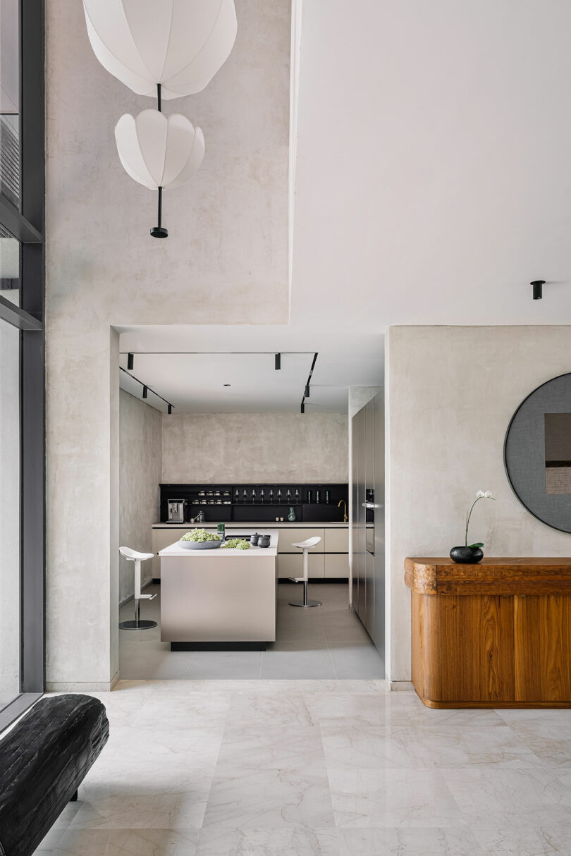 Modern kitchen with sleek beige cabinets, an island, and white pendant lights in a room with concrete walls and marble flooring.