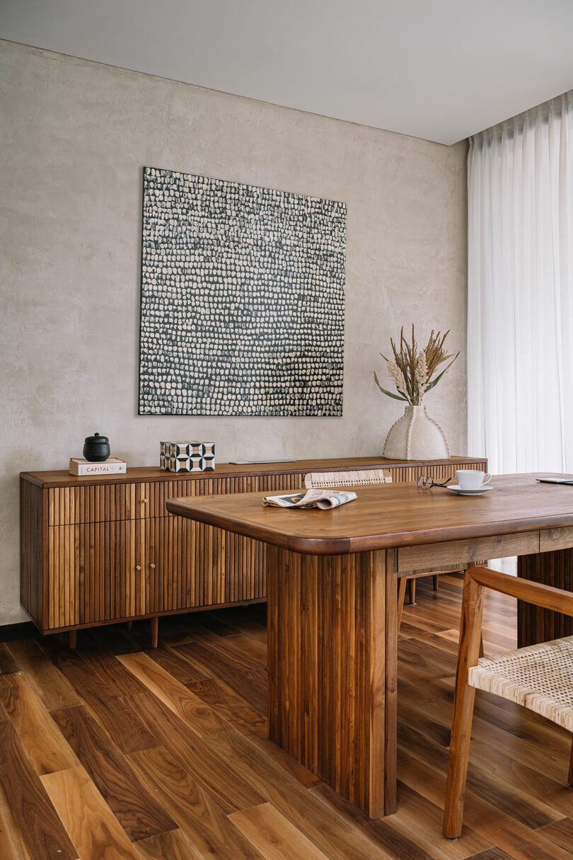 Modern dining room with wooden furniture including a table, chairs, and sideboard. a large abstract painting hangs on the concrete wall, with a vase and decor on the sideboard.