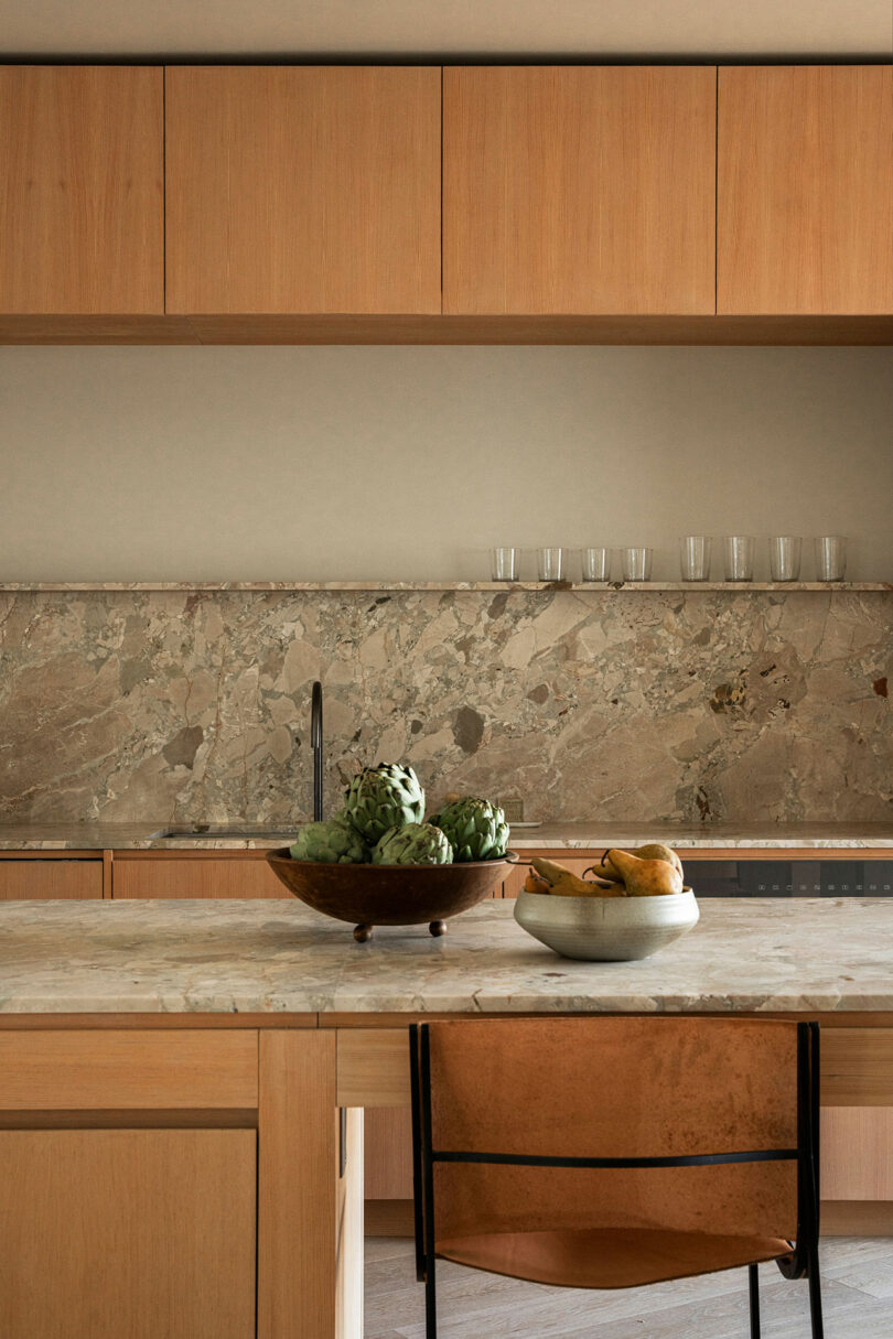 A kitchen with wooden cabinets, a marble countertop, a bowl of artichokes, a bowl of pears, and six drinking glasses on a marble backsplash. A tan leather chair is partially visible.