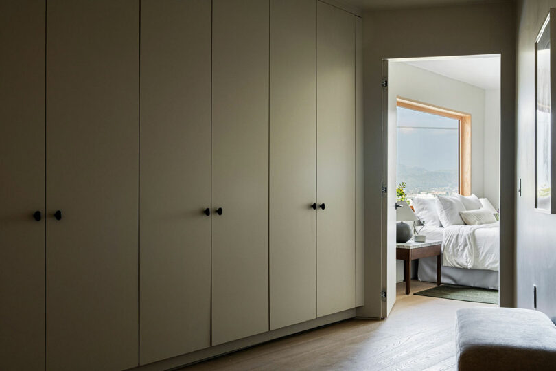 A hallway with closed white closets on the left leads to a bedroom with a bed, pillows, and a large window.