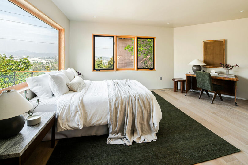 A bedroom with a large window, white bedding, wooden bedside table with a lamp, green rug, and a desk with a chair against a beige wall.