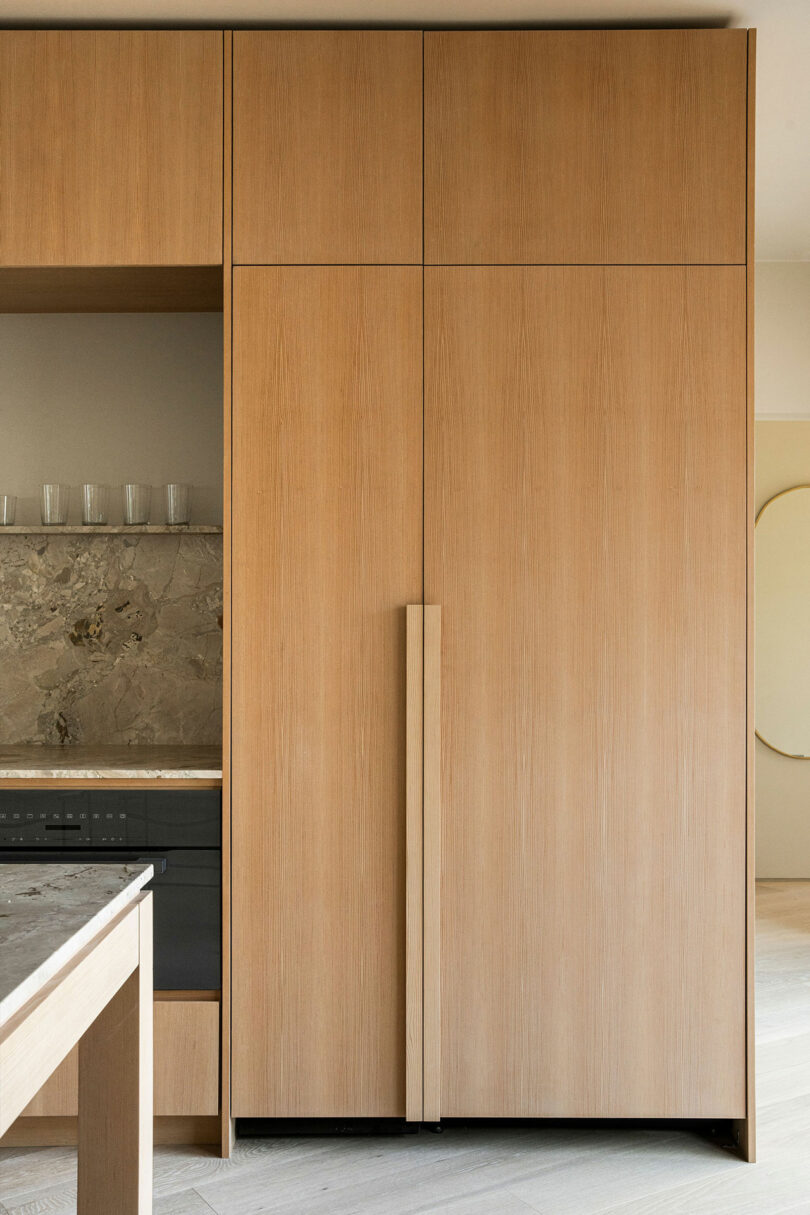 A modern kitchen with light wood cabinets, integrated appliances, a marble backsplash, and a tall, vertical pull handle on the cabinetry.