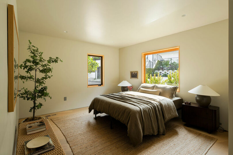 A neatly arranged bedroom with a bed in the center, flanked by two nightstands with lamps. A potted plant stands on the left side. Two windows allow natural light to illuminate the space.