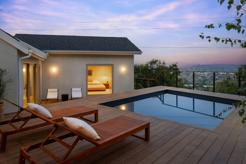 A modern backyard features a wooden deck with two lounge chairs beside a rectangular pool. Sliding glass doors open to a bedroom, with a view of a hilly landscape under a pink and blue sky.