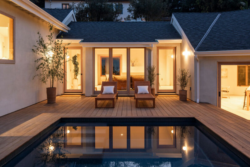 View of the backside of a modern, lit up house at twilight with a pool surrounded by a dark wooden deck.