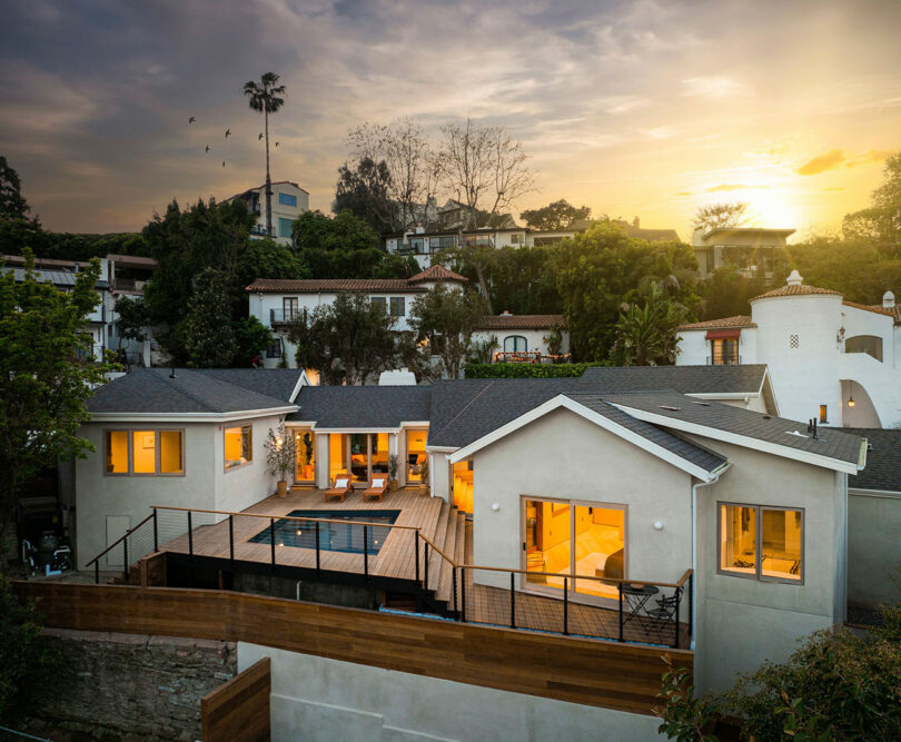 A modern house with large windows and a wooden deck featuring a small pool. The house is situated on a hillside with other houses nearby, and the sun is setting in the background.