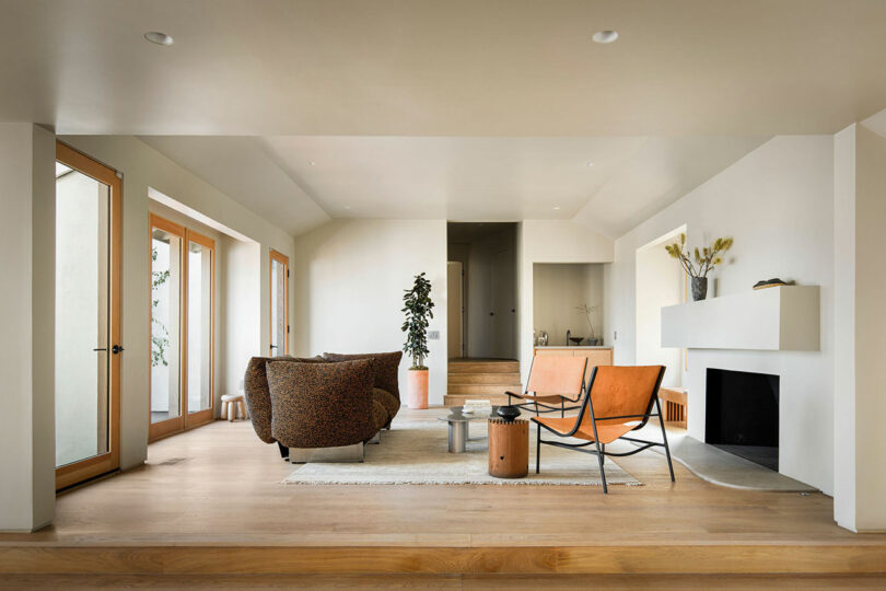 A modern living room with light wood flooring, neutral walls, minimalistic furniture including a sofa, chairs, coffee table, and a fireplace. There is a plant and some decor in the room.