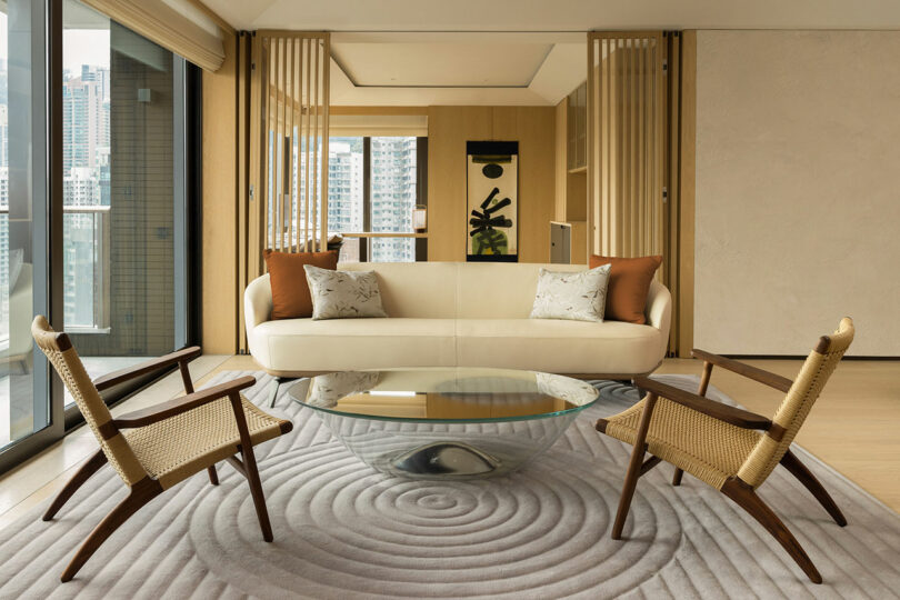 Modern living room interior with a beige sofa, two wooden chairs, a round glass coffee table, and a city view through large windows.