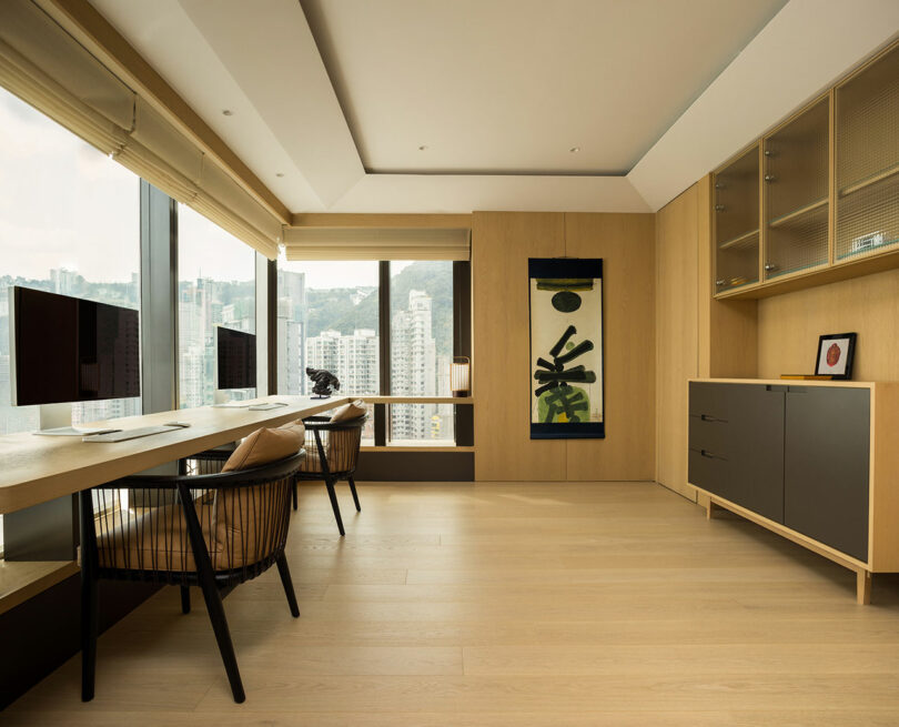 Modern home office with two computer monitors on a wooden desk, floor-to-ceiling windows offering a city view, and minimalist decor.