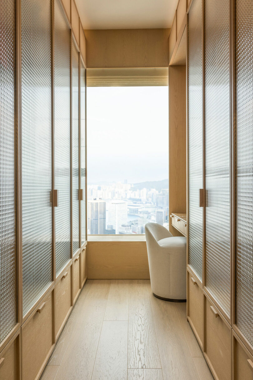 Interior of a modern, narrow room with wooden floors and walls, featuring a single armchair near a large window with a city view, and translucent sliding doors on either side.