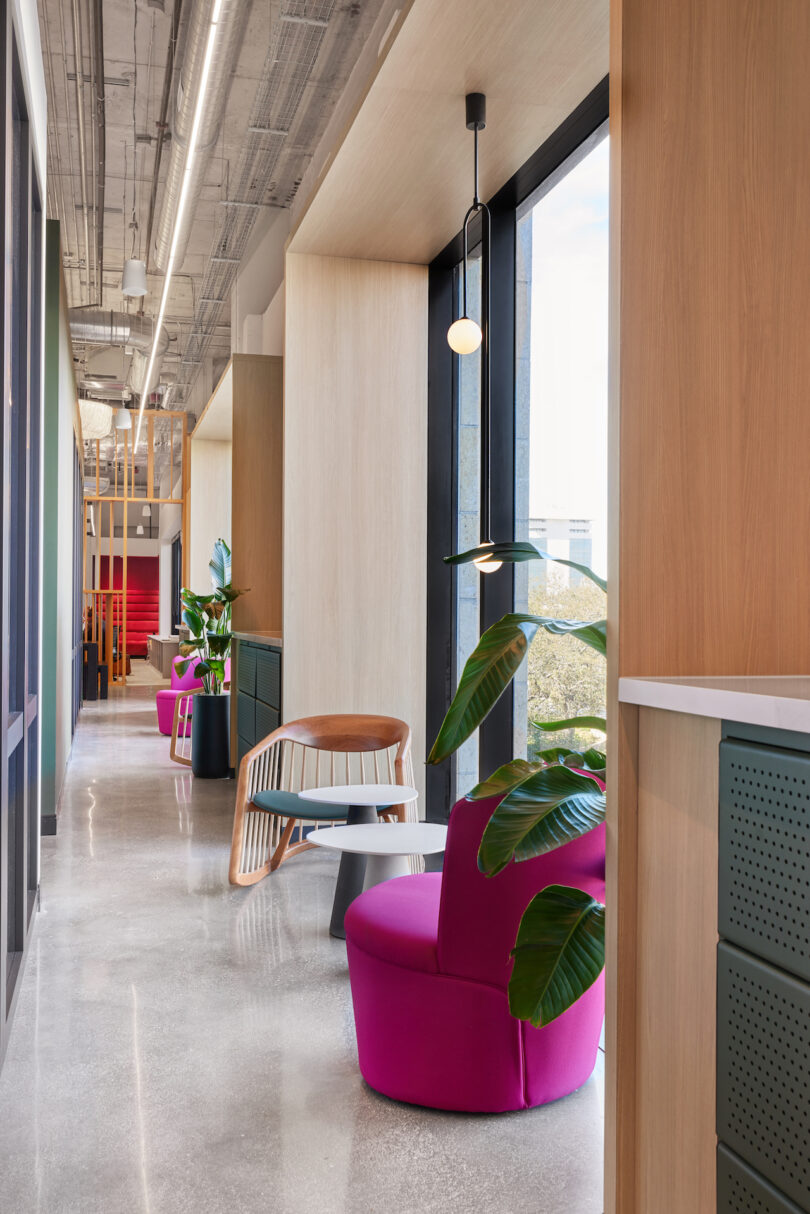 Modern office interior featuring a vibrant pink chair, a wooden chair, hanging lights, and large windows with views of the city outside