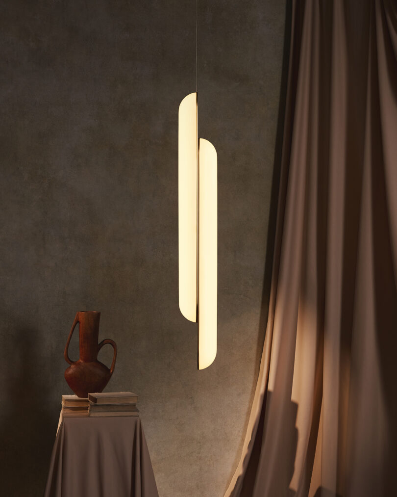 Modern pendant light illuminating a rustic jug on a pedestal, with draped curtains in a dimly lit room with textured walls.