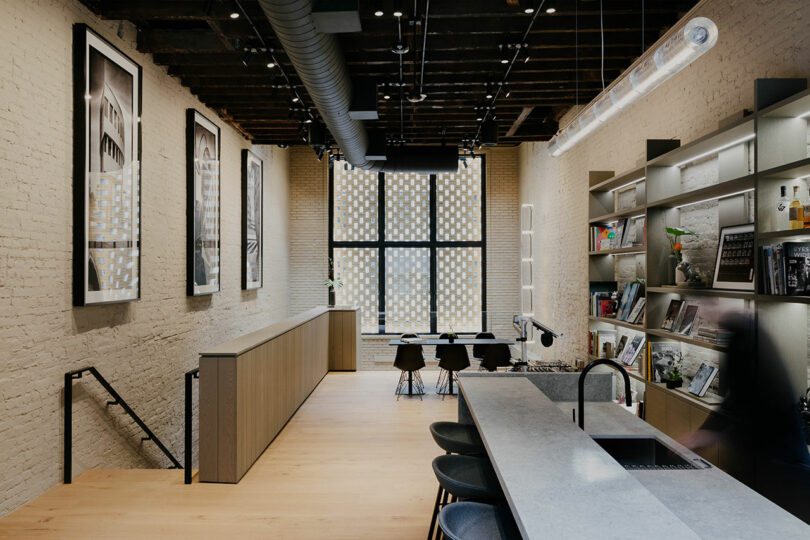 Modern kitchen and dining area with exposed brick walls, wooden floors, a long kitchen island, black stools, and framed artwork on the walls.