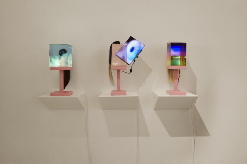 Three digital art displays on pedestals against a beige wall, each screen showing a different abstract colorful image