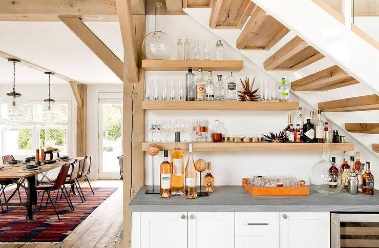 Under staircase kitchen bar design completed with blond wood bar shelves mounted on a white wall over a concrete countertop and white shaker cabinetry.